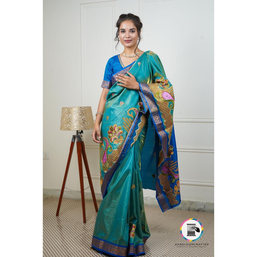 A woman stands elegantly in a peacock blue Banarasi silk saree with intricate gold and pink floral designs, showcasing its beauty and drape.