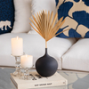 Black ceramic vase with a rounded base and narrow neck, styled with a dried palm leaf in a cozy living room setting.