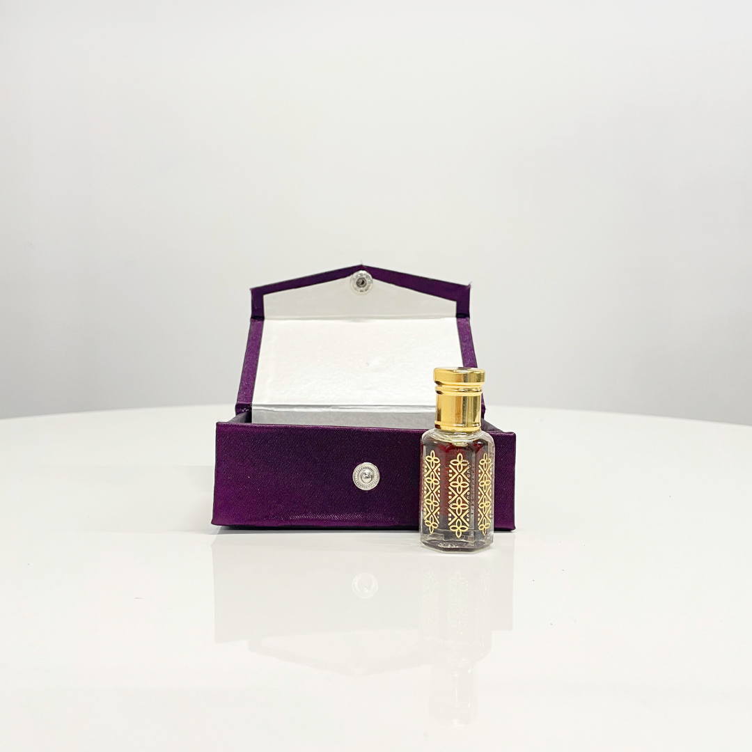 A bottle of Rose Attar Unisex perfume, presented in an elegant purple and gold box, sits on a white table.