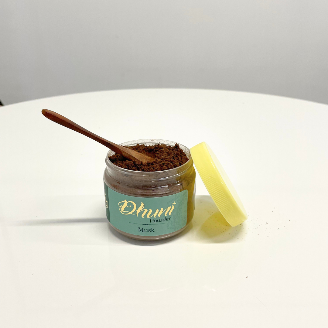 A jar of Dhuni Powder in Musk scent with a small wooden spoon resting inside and the yellow lid placed beside it.