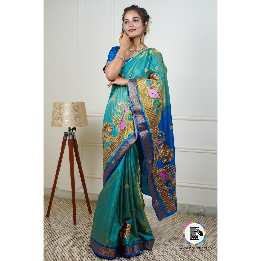 A woman standing wearing a peacock blue Banarasi tissue silk saree with intricate gold and pink paisley embroidery.