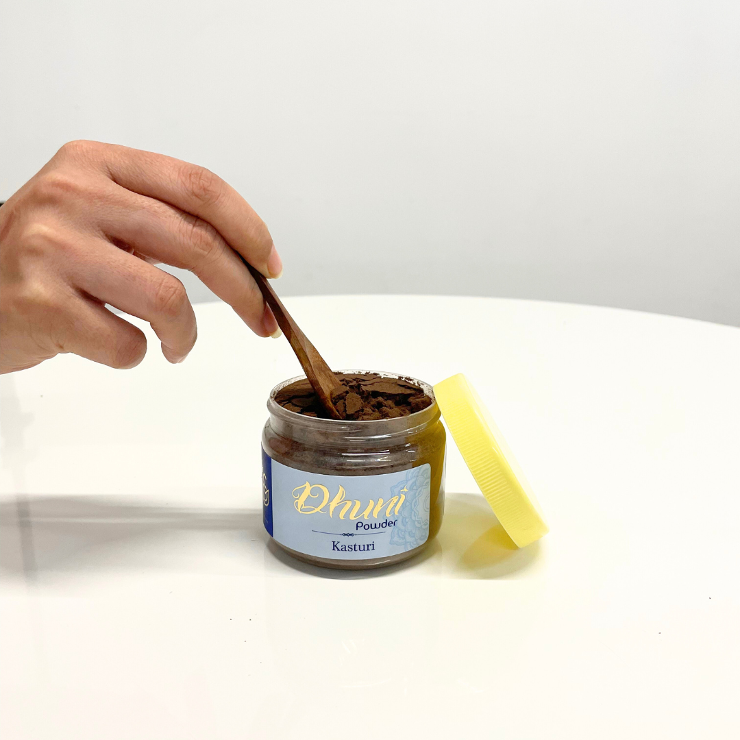 A jar of Kasturi Dhuni Powder with a wooden spoon scooping out the aromatic incense, ready to create a calming atmosphere.