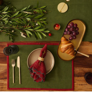 Festive green placemat and napkin set with red border, styled on a holiday table with forest greenery.