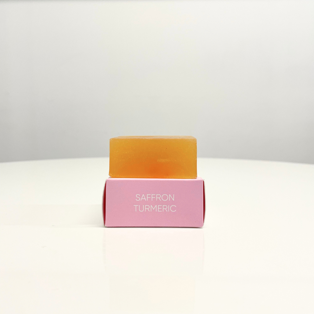 A single bar of golden-hued Saffron Turmeric Handcrafted Soap rests on its pink box.