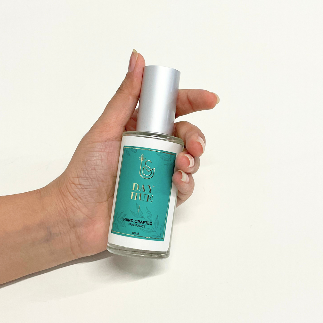 A hand holds a bottle of The Day's Hue Perfume, a unisex fragrance with a teal and gold label.