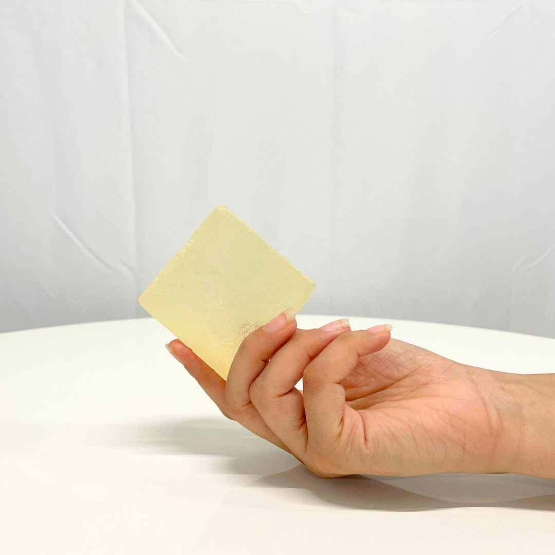 A hand holds a square bar of translucent, light yellow Jasmine Handcrafted Soap.