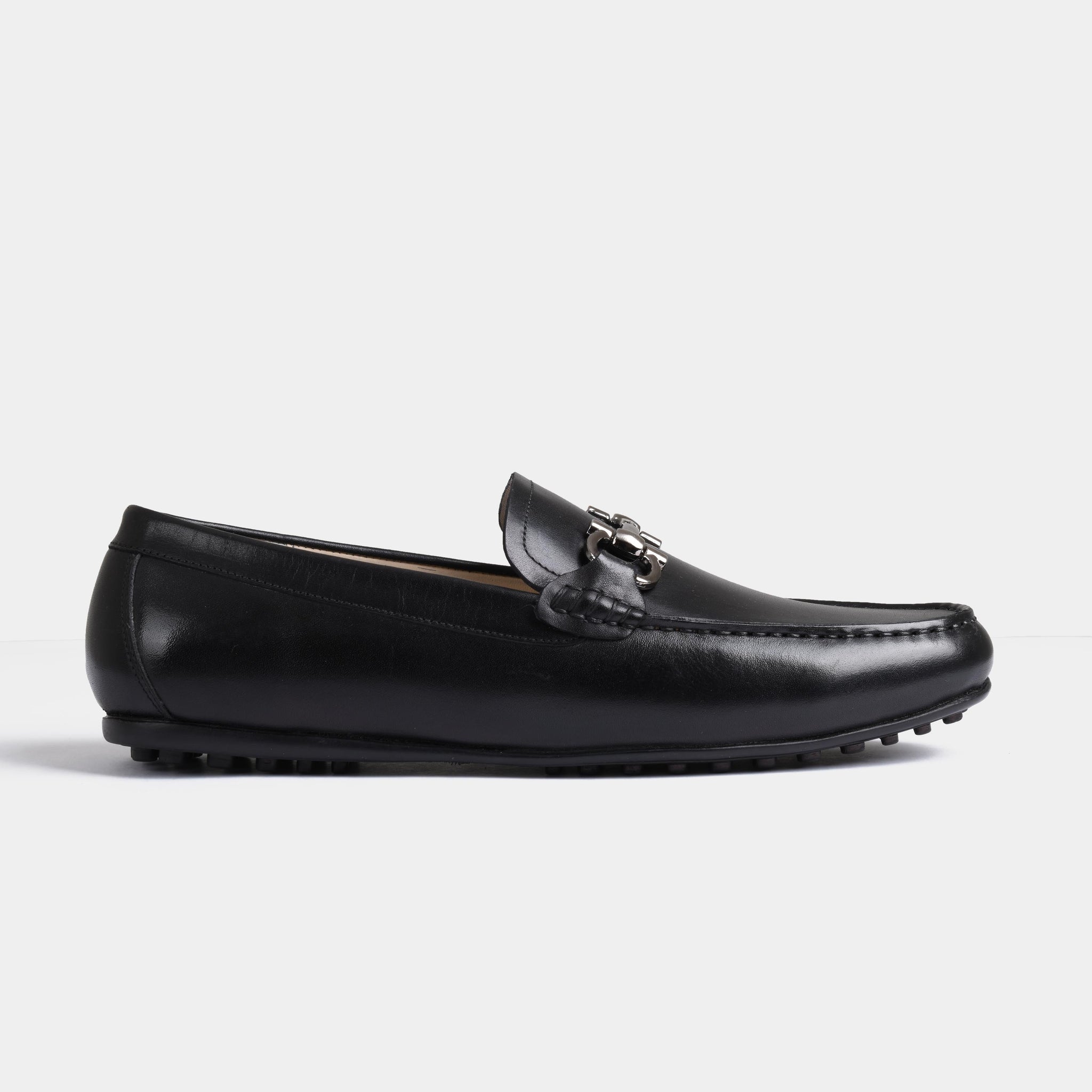 A side view of the Helio Black Leather Driving Loafers showcasing their sleek black leather, metal buckle detail, and classic moccasin stitching.