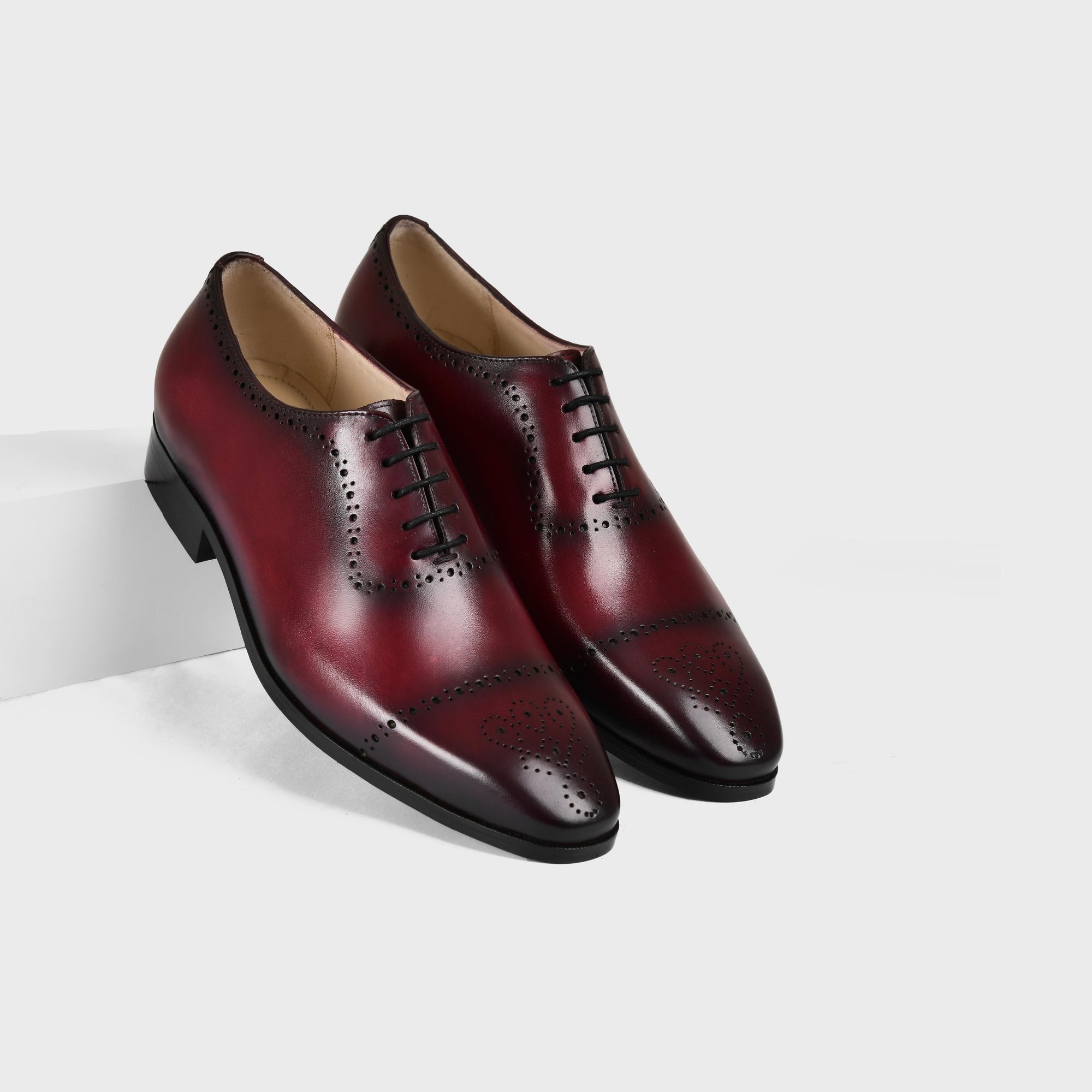 A pair of Lonzo Wine Lace Up shoes, crafted from burgundy leather with decorative broguing, stand on a white surface.