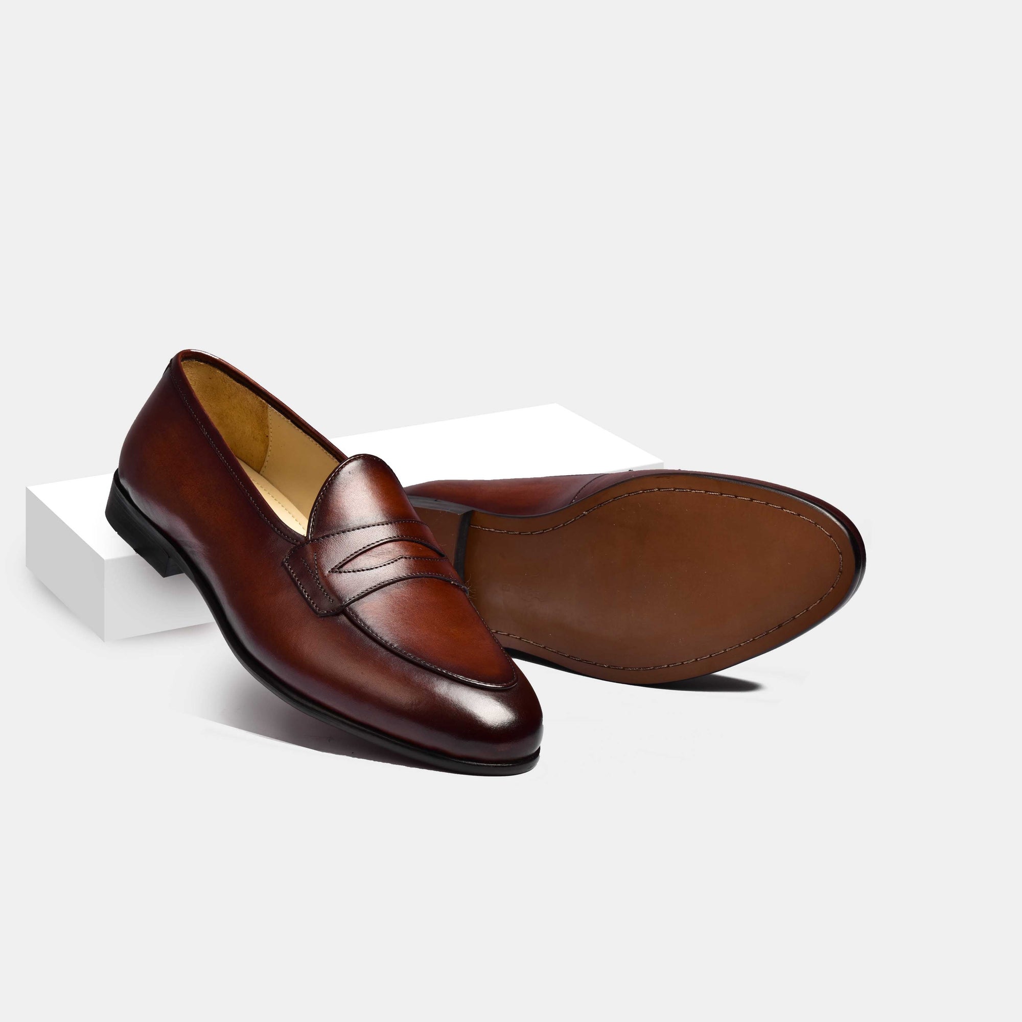 A pair of Arno Loafers in brown patina leather with a grand penny loafer strap, showcased on a white surface.