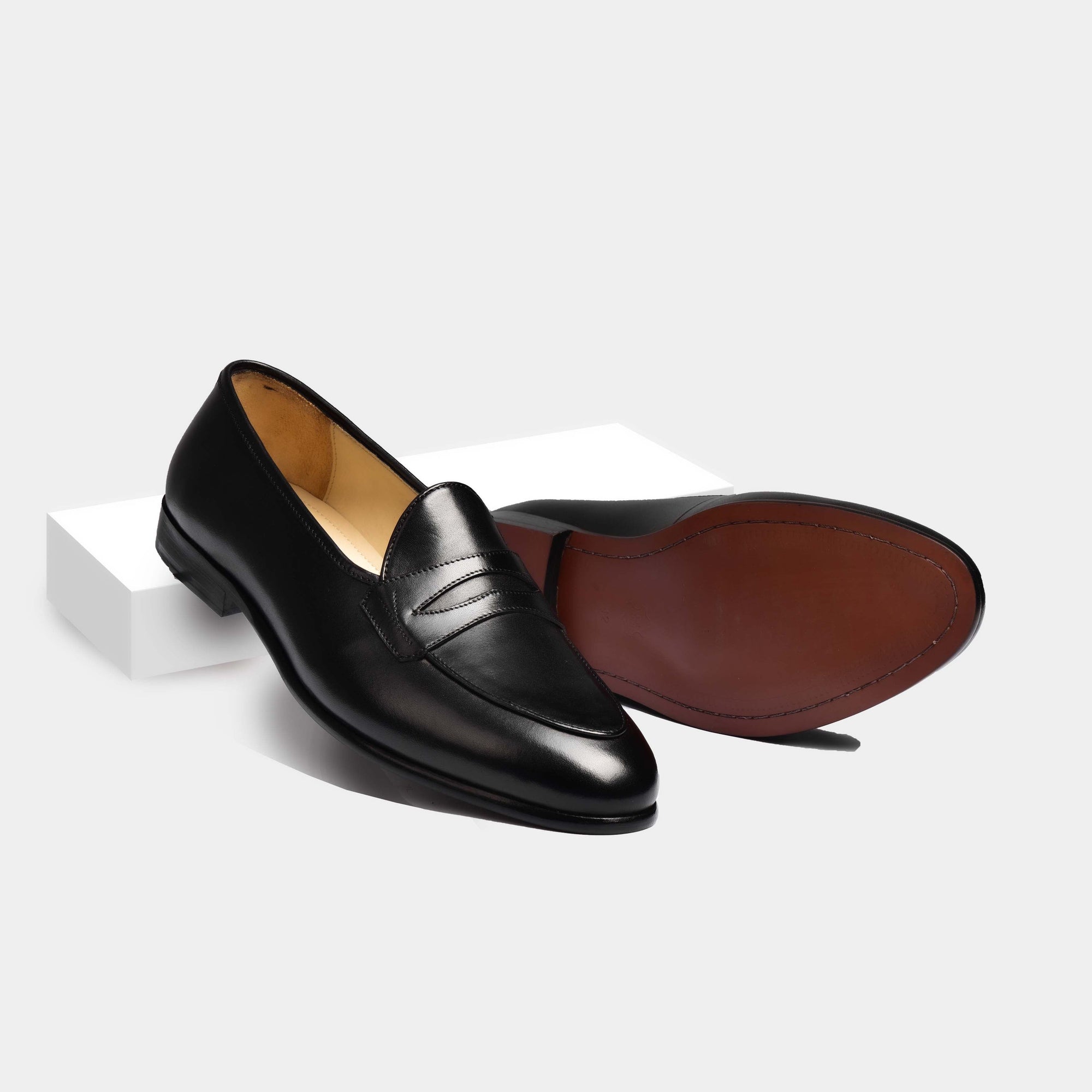 Pair of black patina Arno Loafers with a grand penny loafer strap across the front. One shoe is upright and the other is on its side, revealing the sole.