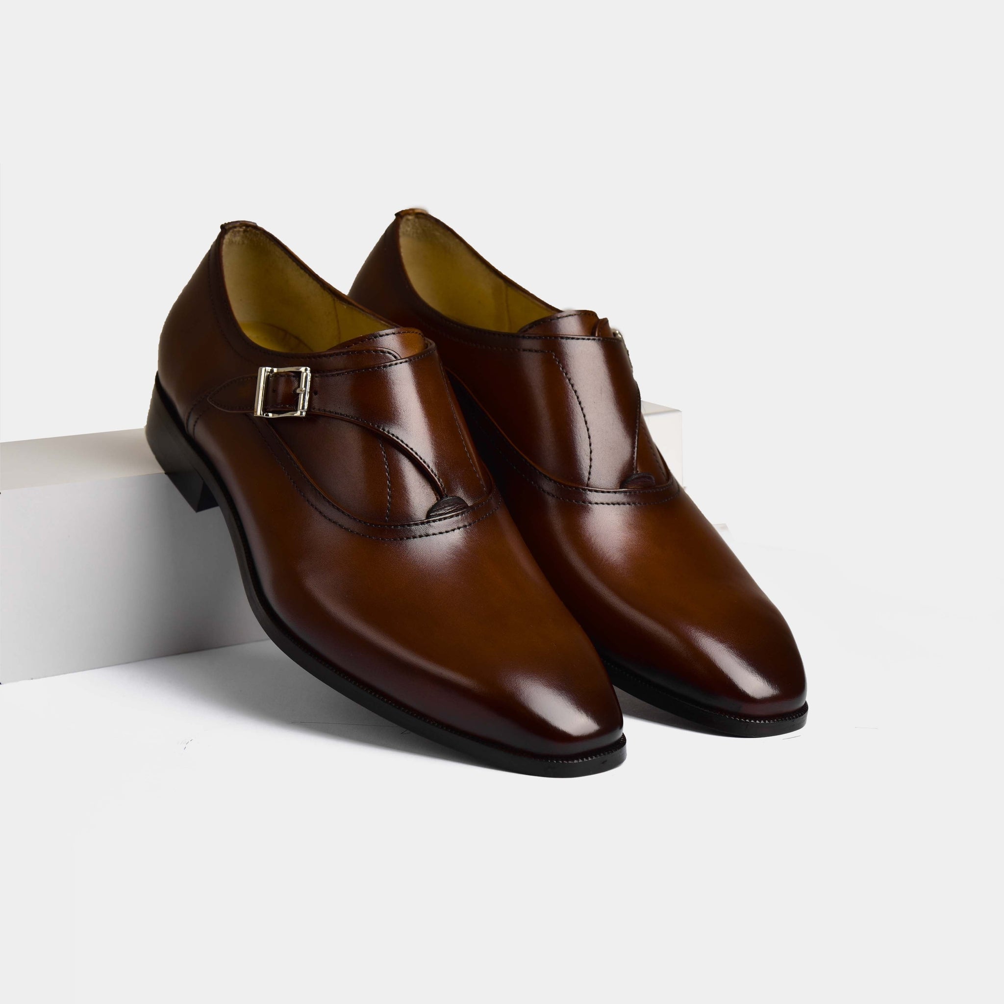 A pair of Efron Choc Monk Straps, crafted from polished brown leather, stand on a white surface.