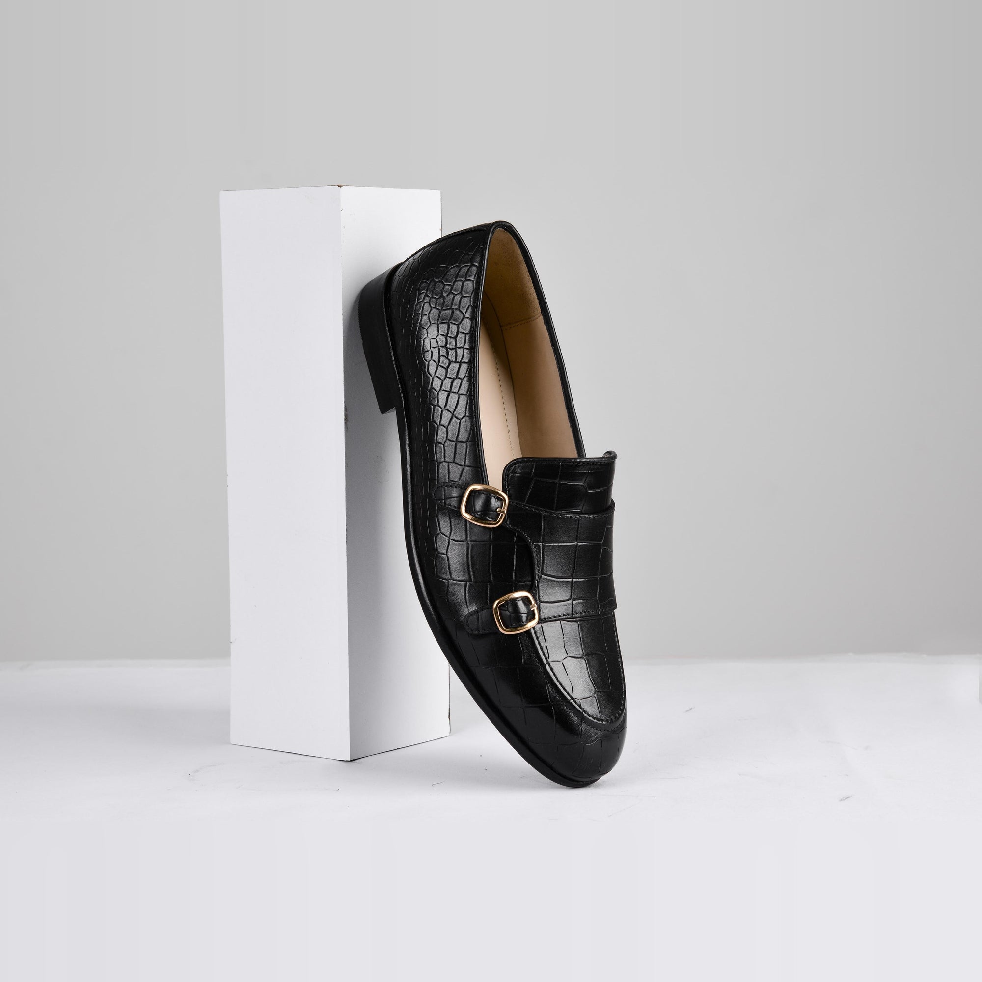 Black leather monk strap shoe with a croc-embossed pattern and gold buckles, resting against a white block.